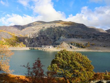 A lake with a bridge and buildings at the base of a rocky mountain with trees in the foreground at 9 Ty Du Road in Llanberis