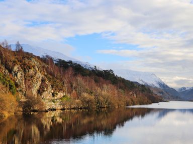 A lake with trees and hills on the shore and snow covered mountains in the background at 9 Ty Du Road in Llanberis
