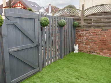 A fenced outdoor area with artificial grass a gray wooden gate three round green shrubs on the fence a small white statue near a brick wall at Sunnydene in Paignton