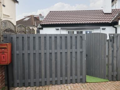 A closed grey wooden fence with a gate and a red post box beside it outside a house with a tiled roof