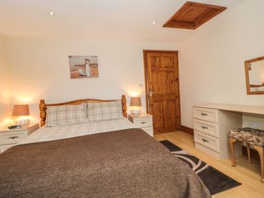 A bedroom with a wooden double bed a brown blanket two bedside tables with lamps a wooden door a desk with drawers a floral chair and a wall painting at Little Walton in Burnham-On-Sea