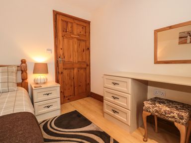 A bedroom with a wooden door a bed with checked bedding a nightstand with a lamp a desk with drawers a patterned stool and a wall mirror at Little Walton in Burnham-On-Sea