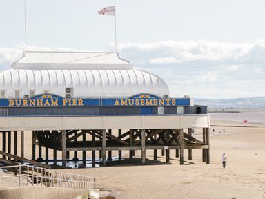 A pier with a building labeled Burnham Pier Amusements on a sandy beach with a few people walking at Little Walton in Burnham-On-Sea