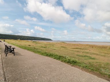 A paved walkway with benches beside a grassy area near a beach with hills in the distance at Little Walton in Burnham-On-Sea