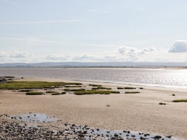 A coastal landscape with a sandy beach rock pools patches of grass water and hills in the background at Little Walton in Burnham-On-Sea