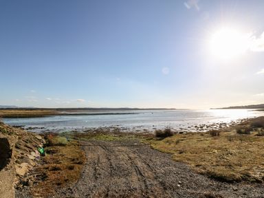 A shoreline with gravel path leading to water under a clear sky with the sun shining at Malltraeth Cottage in Malltraeth