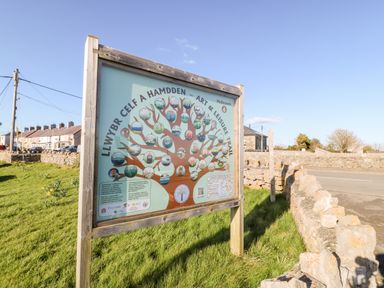 An outdoor wooden sign with a tree diagram and text about an art and leisure trail on grass near a stone wall in a residential area at Malltraeth Cottage in Malltraeth