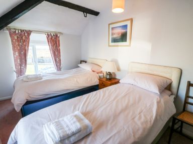 A bedroom with two single beds a nightstand with a lamp a framed picture and pink curtains at Malltraeth Cottage in Malltraeth
