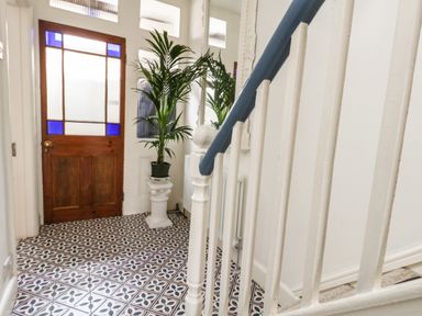 A hallway with patterned tile floor a wooden door with blue stained glass and a potted plant on a white pedestal at ODYSEA in Weymouth