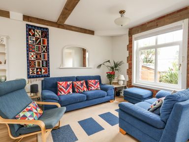 A living room with blue sofas and armchair with patterned cushions a wall hanging and a large window showing plants outside at ODYSEA in Weymouth