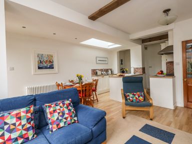 A living area with a blue sofa and chair with colorful cushions a wooden dining table set with dishes a kitchen area with white cabinets and a wooden door at ODYSEA in Weymouth