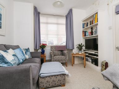 A living room with a grey sofa and blue pillows a patterned armchair a small wooden table with a plant a television on a built-in shelf and a window with purple curtains at ODYSEA Weymouth