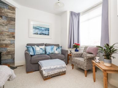 A sitting room with a grey sofa with blue cushions a patterned armchair with a pink cushion an ottoman with a throw a wooden side table with a plant and a mug and a window with curtains at ODYSEA in Weymouth