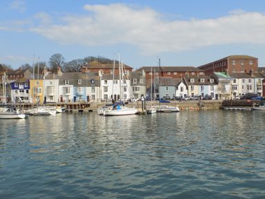A harbor with sailboats docked in front of a row of colorful houses and brick buildings at ODYSEA in Weymouth