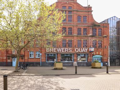 A brick building with a sign reading Brewers Quay a tree benches bicycles and Weymouth Museum entrance at ODYSEA in Weymouth