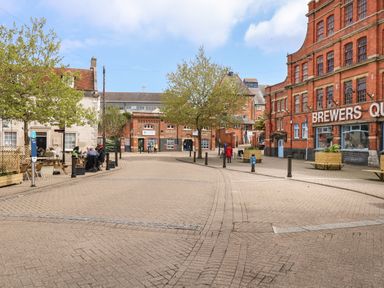 A public square with brick buildings trees benches and people walking at ODYSEA in Weymouth