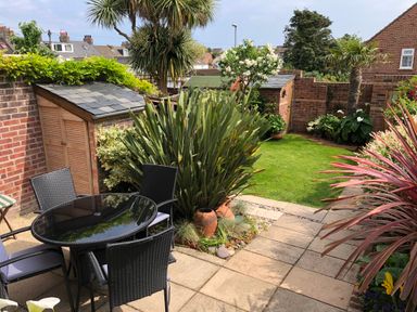A patio with a glass table and wicker chairs next to a garden with plants and shrubs at ODYSEA in Weymouth