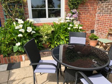 An outdoor patio with a glass table and four wicker chairs with cushions next to a brick wall with a window and white flowers at ODYSEA Weymouth