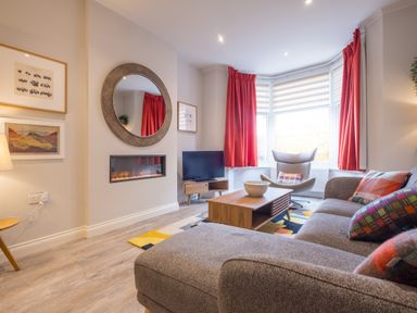 A living room with a gray sofa, wooden coffee table, armchair by a bay window with red curtains at Howe Keld 1 Ground floor Keswick