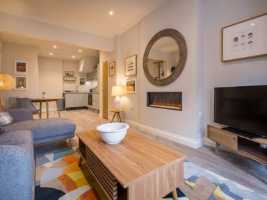 A living room with a gray sofa wooden coffee table a wall-mounted electric fireplace and an open kitchen area at Howe Keld 1 Ground floor in Keswick