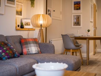 A living room with a gray sofa with patterned cushions a wooden floor lamp and a dining table with chairs at Howe Keld 1 Ground floor in Keswick
