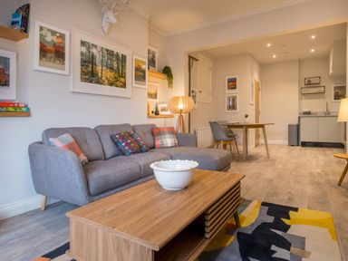 A living room with a grey sofa and colorful cushions a wooden coffee table a multi-colored rug framed pictures on the wall a dining table with chairs and a small kitchen area at Howe Keld 1 Ground floor in Keswick