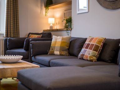 A living room with dark sofas, patterned cushions, a wooden coffee table, a lamp, plants, and curtains at Howe Keld 4 First floor in Keswick