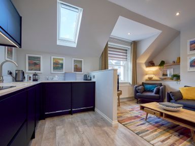 A kitchen area with dark cabinets and a living room with a sofa armchair coffee table and wall shelves at Howe Keld 4 First floor in Keswick
