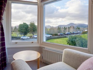 A corner seating area with a round wooden table and two chairs near windows showing a street with parked cars and trees at Howe Keld 4 First floor in Keswick
