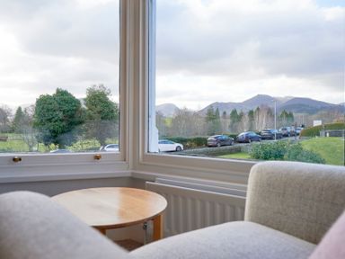 A small round wooden table and part of a chair by a large window showing parked cars trees and mountains outside at Howe Keld 4 First floor in Keswick