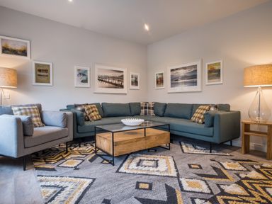 A living room with a sectional sofa armchair coffee table lamps and framed pictures on the wall at Howe Keld 5 First Floor in Keswick