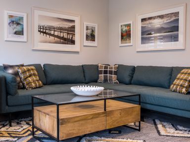 A corner living room with a blue sectional sofa with cushions a glass and wood coffee table and five framed pictures on the walls at Howe Keld 5 First Floor in Keswick