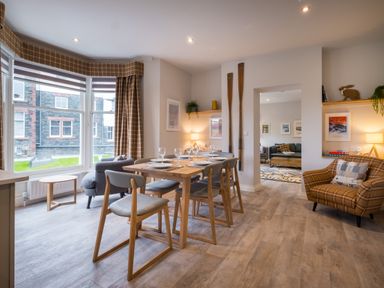 A dining area with a wooden table set for six by a large window and an adjacent living area with armchairs at Howe Keld 5 First Floor Keswick