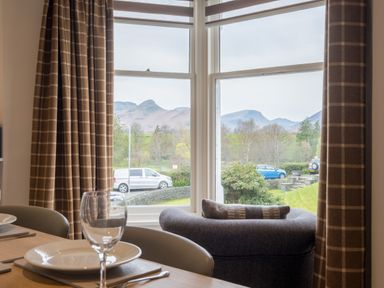A dining area with plates and glasses on a table next to windows with brown check curtains and a view of cars and mountains at Howe Keld 5 First Floor in Keswick