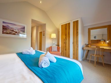 A bedroom with a bed covered in a blue blanket and rolled towels a wooden wardrobe an orange armchair a desk with a chair and a large mirror at Howe Keld 5 First Floor in Keswick