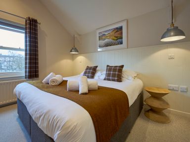 A bedroom with a double bed with brown plaid pillows and white towels on the bed a wooden side table and pendant lights Howe Keld 5 First Floor Keswick