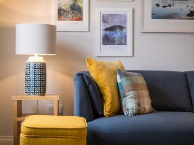 A living room with a blue sofa with two cushions a yellow ottoman a wooden side table with a patterned lamp and three framed pictures on the wall at Howe Keld 6 Second Floor in Keswick