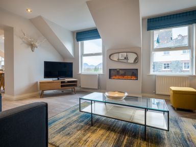 A living room with a glass coffee table a TV on a wooden stand a wall-mounted electric fireplace and a yellow ottoman at Howe Keld 6 Second Floor in Keswick