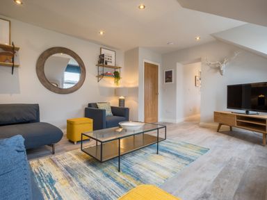 A living room with a blue sofa an armchair a glass coffee table a yellow ottoman a wooden TV stand a wall mirror and shelves at Howe Keld 6 Second Floor in Keswick