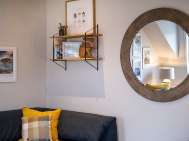 A corner of a room with a navy sofa and patterned cushions a wall shelf with books and decorations and a round wooden framed mirror on the wall at Howe Keld 6 Second Floor in Keswick