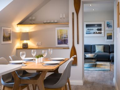 A dining area with a wooden table set with plates and glasses and a living room with a sofa and framed pictures at Howe Keld 6 Second Floor in Keswick