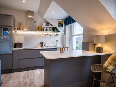 A kitchen with gray cabinets a white countertop a row of hanging mugs a window with a blue blind and a lamp on the counter at Howe Keld 6 Second Floor in Keswick