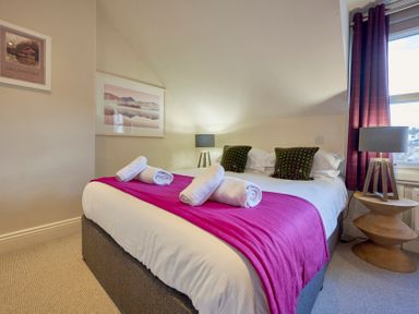 A bedroom with a double bed white bedding purple blanket and rolled towels two bedside tables with lamps at Howe Keld 6 Second Floor Keswick