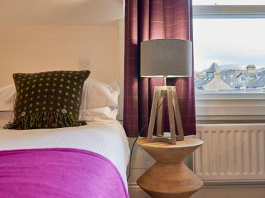 A bedroom with a bed a polka dot pillow a wooden side table and a lamp near a window with a view of rooftops at Howe Keld 6 Second Floor in Keswick