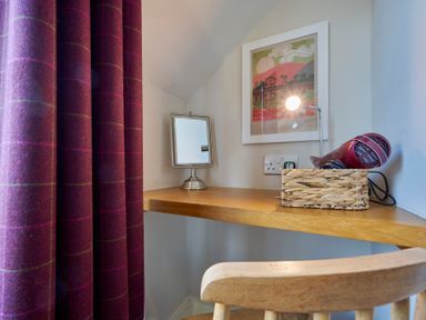 A small wooden desk with a mirror a framed picture a lamp and a hairdryer in a basket by a wooden chair and purple curtains at Howe Keld 6 Second Floor in Keswick