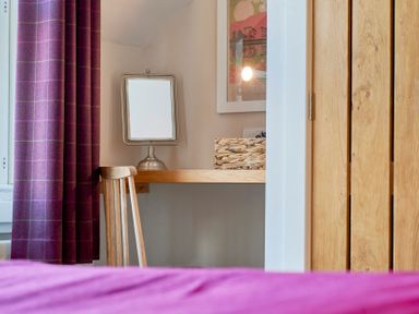 A wooden chair next to a wall-mounted wooden desk with a tabletop mirror and a woven basket near a wooden closet and a window with purple curtains Howe Keld 6 Second Floor Keswick