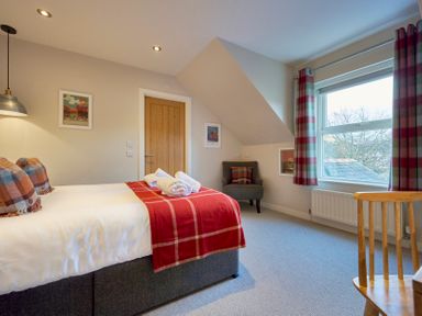 A bedroom with a bed and red plaid blanket a wooden door a window with plaid curtains a grey armchair and a wooden chair at Howe Keld 6 Second Floor in Keswick