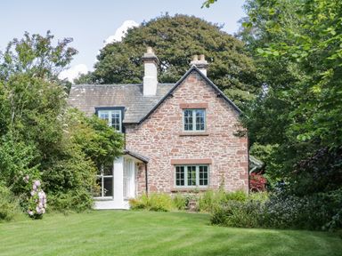 A stone house with green windows surrounded by trees and a lawn at Caldhu Cottage in Holmrook