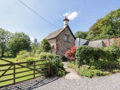 A stone cottage with a chimney surrounded by bushes and trees behind a wooden gate at Caldhu Cottage in Holmrook