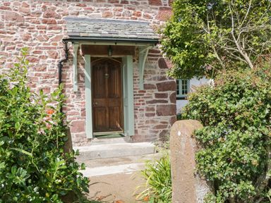 Entrance door with stone steps and a small roof surrounded by bushes and plants at Caldhu Cottage in Holmrook